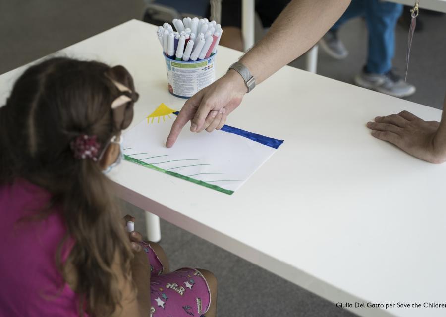 Bambina con codini seduta presa di spalle davanti a un tavolo bianco e davanti a lei c è il braccio di un adulta che indica un foglio sul tavolo