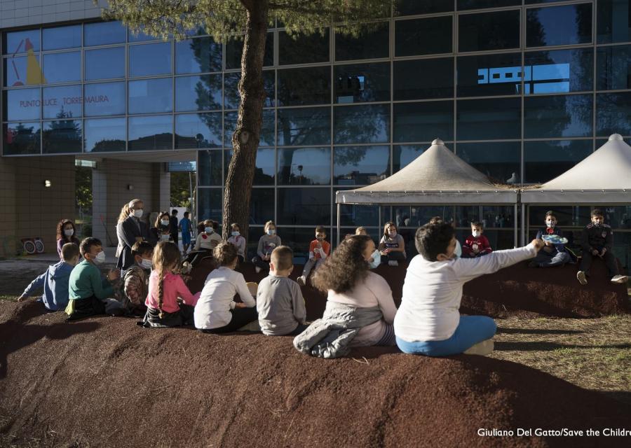 Foto all aperto, una fila di bambini di spalle seduti su un terrapieno guardano due gazebo bianchi montati di fronte a loro