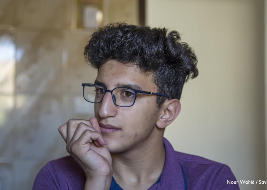 Primo piano di un ragazzo adolescenti con occhiali, capelli neri e ricci abbastanza lunghi. Guarda alla sua sinistra e si tiene il mento con la mano