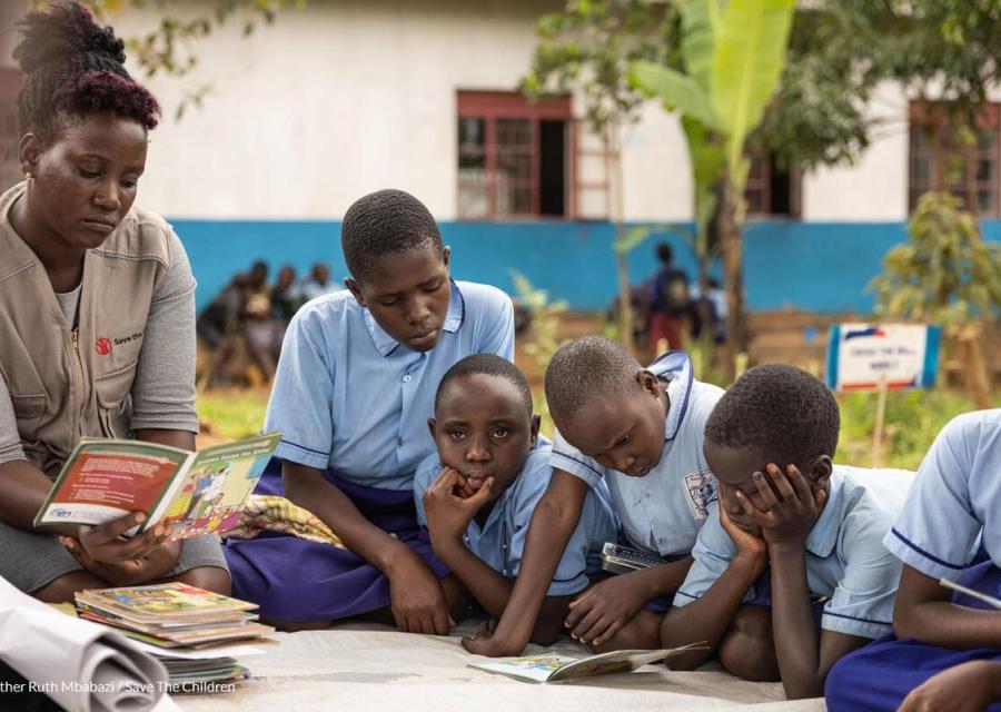 Bambini e bambine in cerchio che leggono un libro accanto all'operatrice di Save the Children 