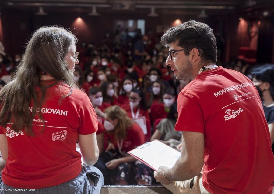 Un ragazzo e una ragazza con la maglietta rossa del movimento giovani di Save the Children discutono su un palco di fronte ad una platea di giovani