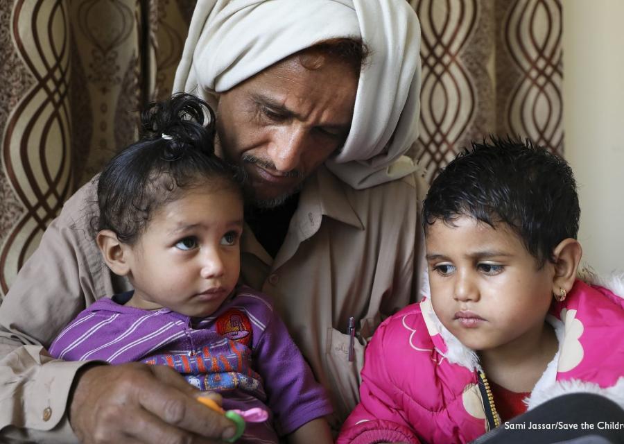 Un papà yemenita con turbante bianco in testa tiene in braccio le sue due bambine e le guarda. Entrambe hanno i capelli corti e neri, una guarda verso l alto e l altra verso la sua sorellina. Indossano magliette viola e rosa