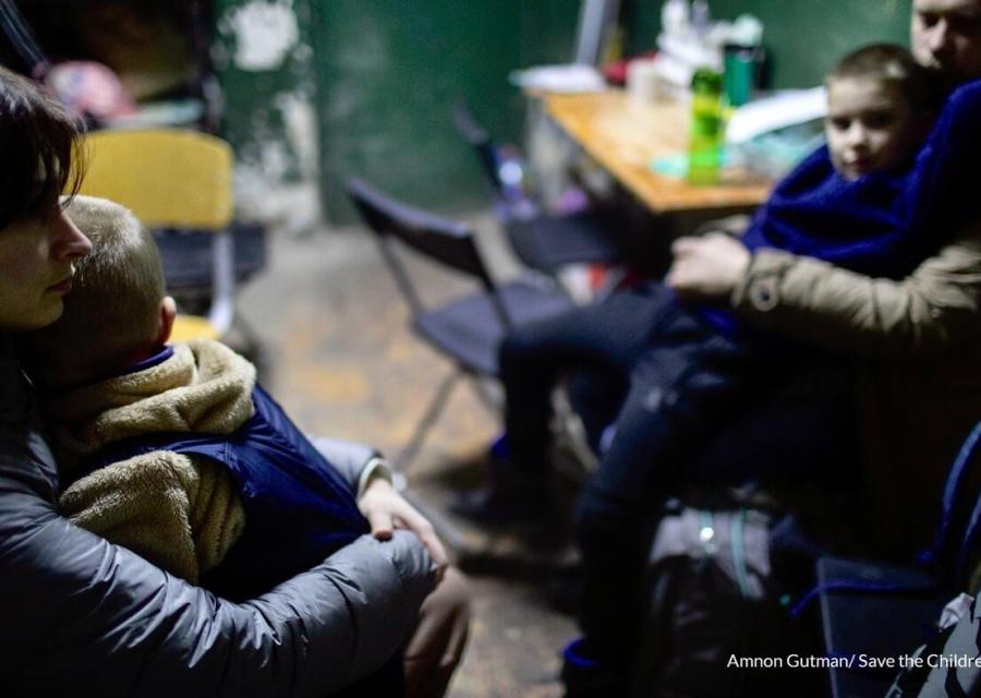 Una mamma ucraina di profilo tiene in braccio il suo bambino da seduta all interno di una stanza con altre persone sedute.