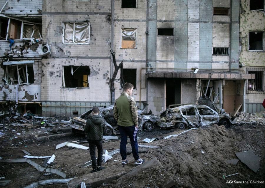 Ucraina, un bambino e un ragazzo in piedi di spalle davanti a edifici e macchine distrutti e in macerie a causa dei bombardamenti