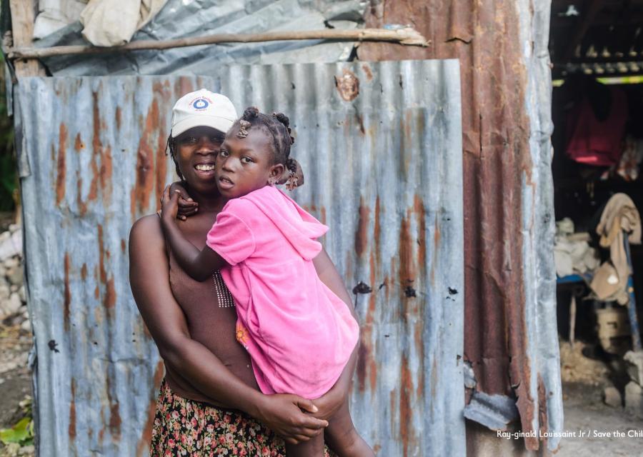 Una donna haitiana tiene in braccio la sua bambina che indossa un vestito rosa e le tiene le braccia strette intorno al collo. La donna indossa una maglietta marrone e un cappellino bianco in testa. Dietro di loro si vedono le lamiere di una baracca.