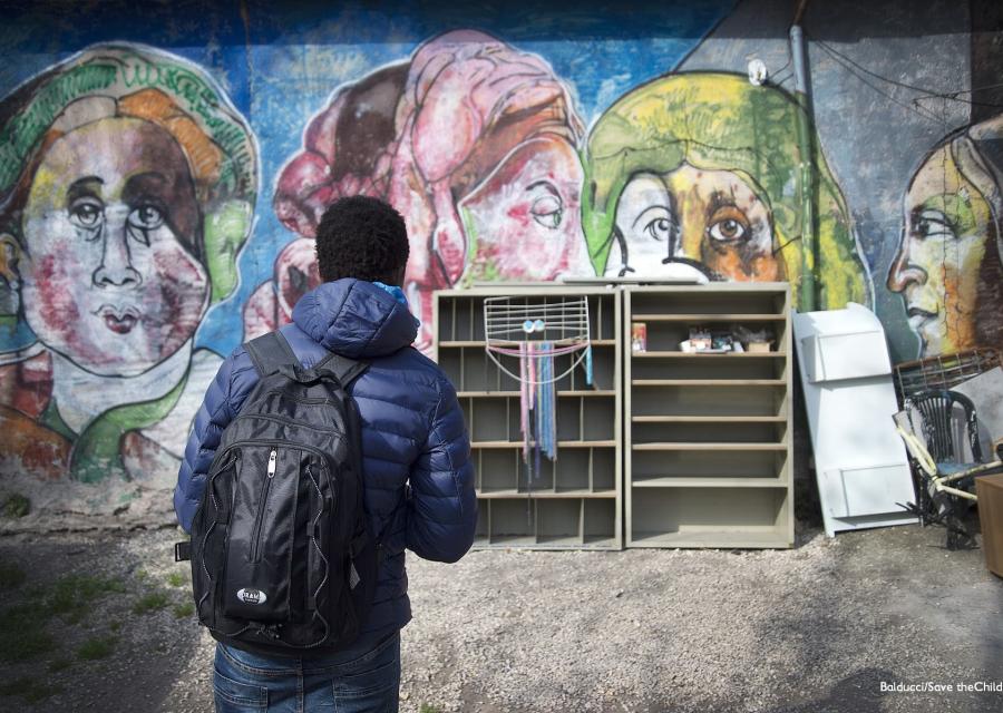 Adolescente di spalle che indossa una giacca pesante blu e tiene sulle spalle uno zaino nero guarda un muro davanti a lui dipinto con un murales e sul muro è appoggiato un mobile con mensole molto rovinato