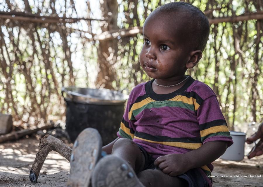 Bambino africano seduto a terra riparato in una capanna indossa una maglietta viola a strisce gialle e guarda alla sua destra.