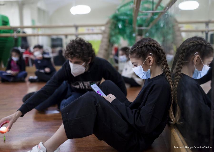 una ragazza e un ragazzo vestiti in tuta nera seduti per terra in un aula di danza, indossano mascherina chirurgica per covid-19, lei legge lui gioca con alcuni giochini per terra