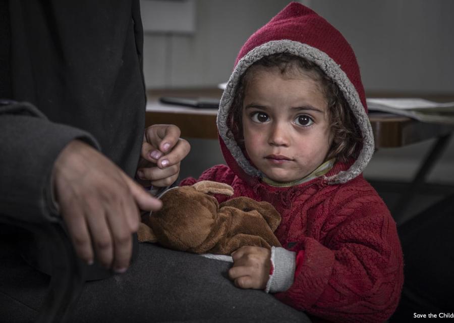 primo piano bambina siriana con cappotto e cappuccio rosso in testa stringe orsacchiotto appoggiata alla gamba di un adulto