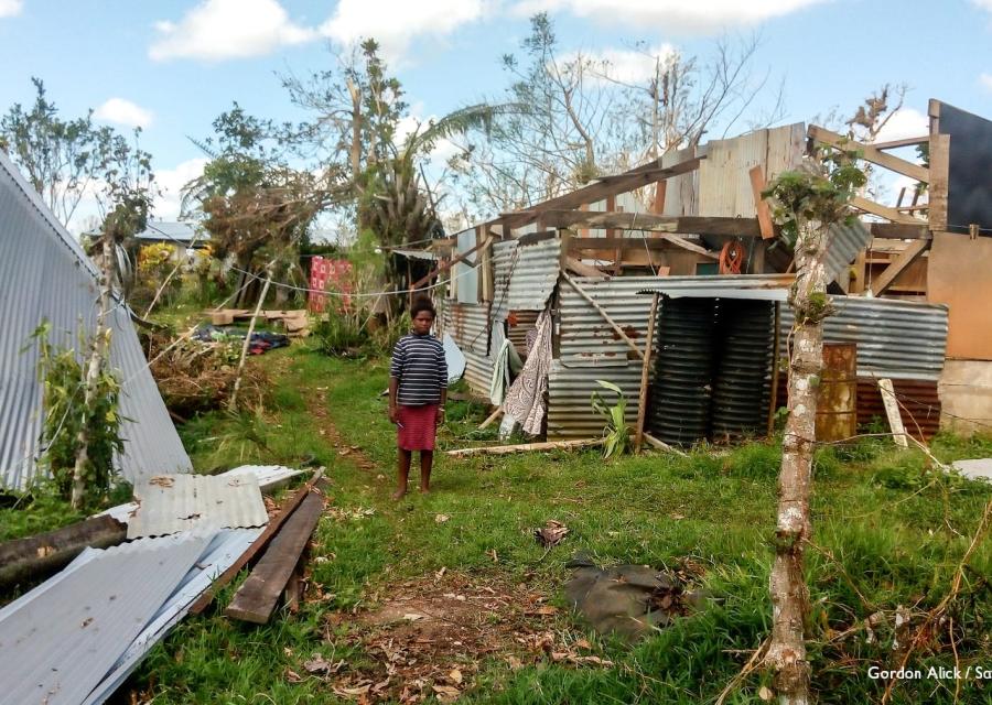 Una bambina in lontananza con gonna rossa e maglietta a righe bianche e nere si trova in piedi e scalza sul prato. Dietro di lei una casa distrutta al passaggio di un ciclone sulle isole Fiji