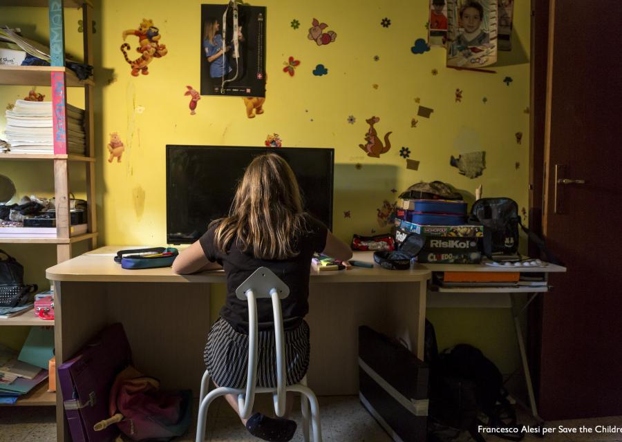 ragazza di spalle seduta alla scrivania della sua cameretta davanti al pc. Intorno a lei giochi da tavola, libri e quaderni.
