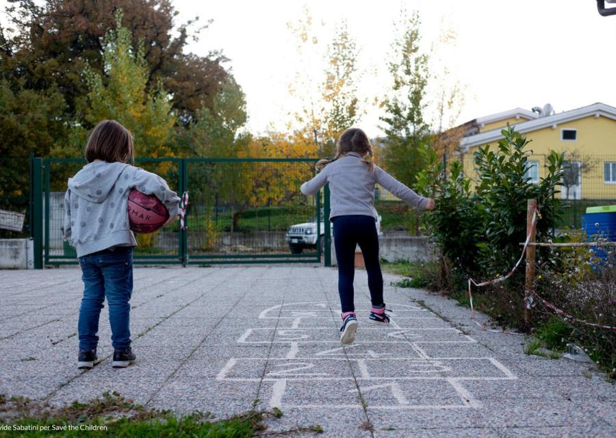 una bambina sulla sinistra tiene un pallone sotto il braccio mentre guarda l'amica che salta sui riquadri a terra del gioco campana