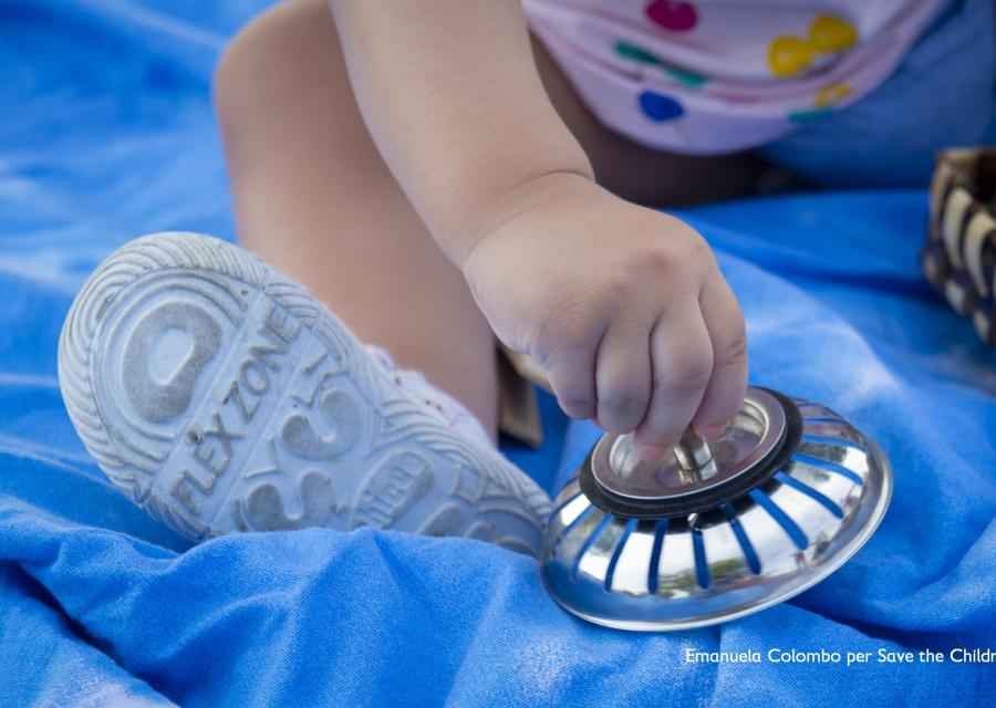 bambina molto piccola seduta su un telo blu gioca con un oggetto che tiene in mano. Nell inquadratura si vedono solo la mano e il piedino