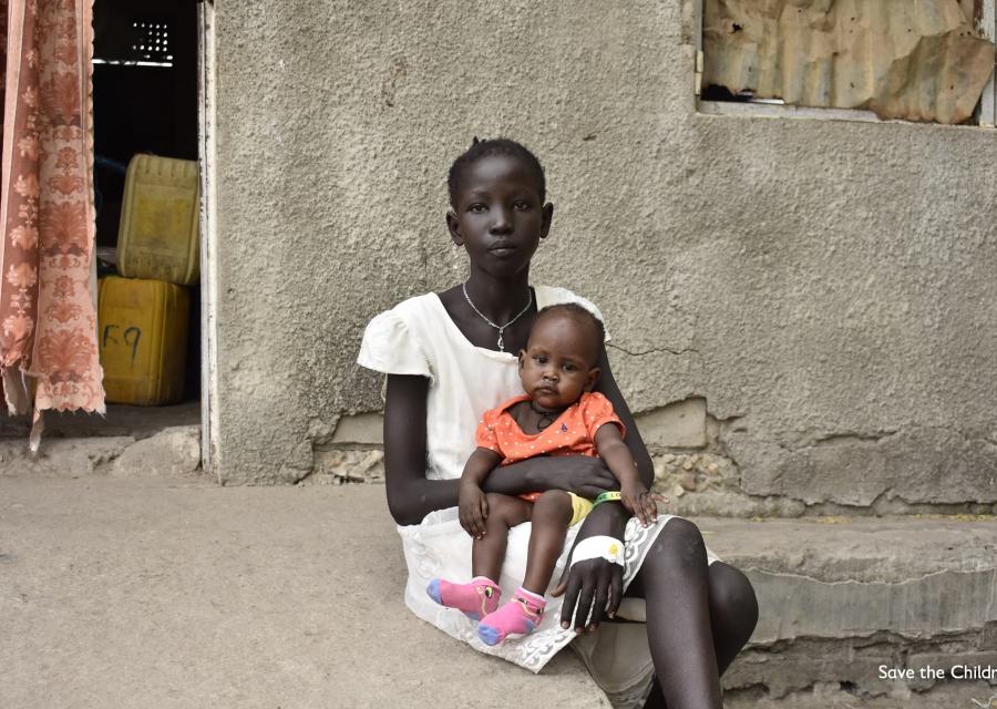 Una ragazzina sud sudanese seduta sui gradini davanti casa sua tiene in braccio una bambina piccola vestita di rosso.