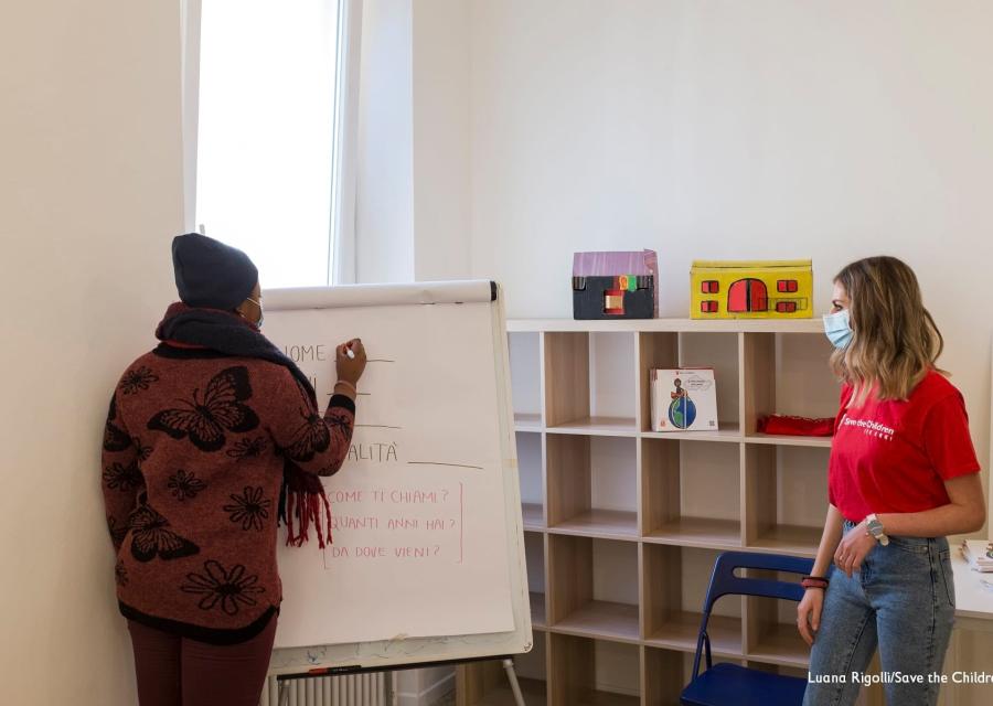 Una ragazza di spalle scrive su una lavagna e un operatrice di Save the Children la guarda all interno del nuovo CivicoZero di Torino