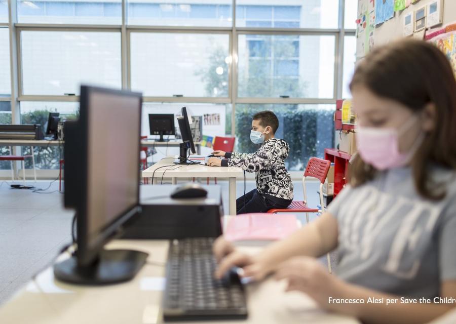 Una bambina di profilo in primo piano sfocata scrive al pc ed e seduta a un tavolo. Al suo lato si vede un altro bambino seduto alla scrivania intenti a scrivere al pc.