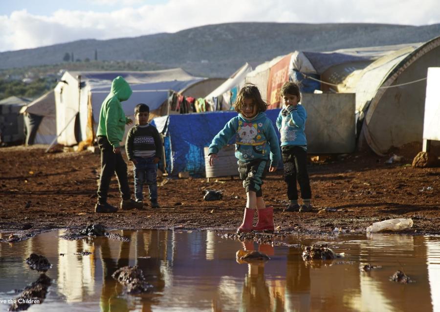 Un gruppo di bambini siriani, quattro in tutto, sono ripresi in piedi da lontano all interno di un campo profughi e su un terreno infangato a causa delle piogge. In particolare, una bambina con stivaletti rosa avanza camminando verso una grande pozzanghera d acqua di fronte a lei.