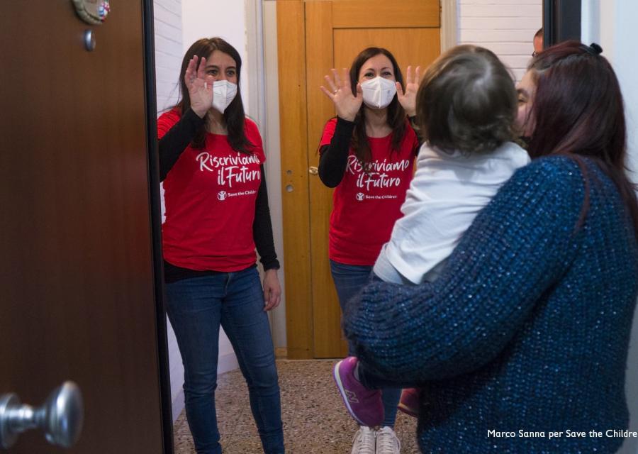 Due operatrici di Save the Children con maglietta della campagna Riscriviamo il Futuro salutano mamma e figlia davanti a loro sull uscio della porta di casa