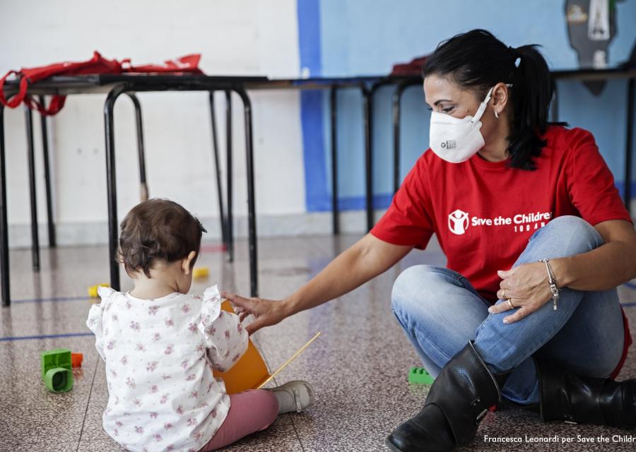 operatrice save the children con maglietta rossa con logo seduta per terra con una bimba di spalle mentre giocano insieme