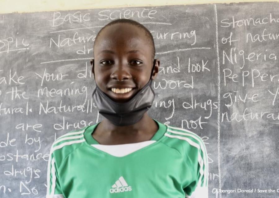 Bambino in primo piano sorridente con mascherina nera abbassata davanti a una  lavagna scritta. Indossa una maglietta verde dell adidas