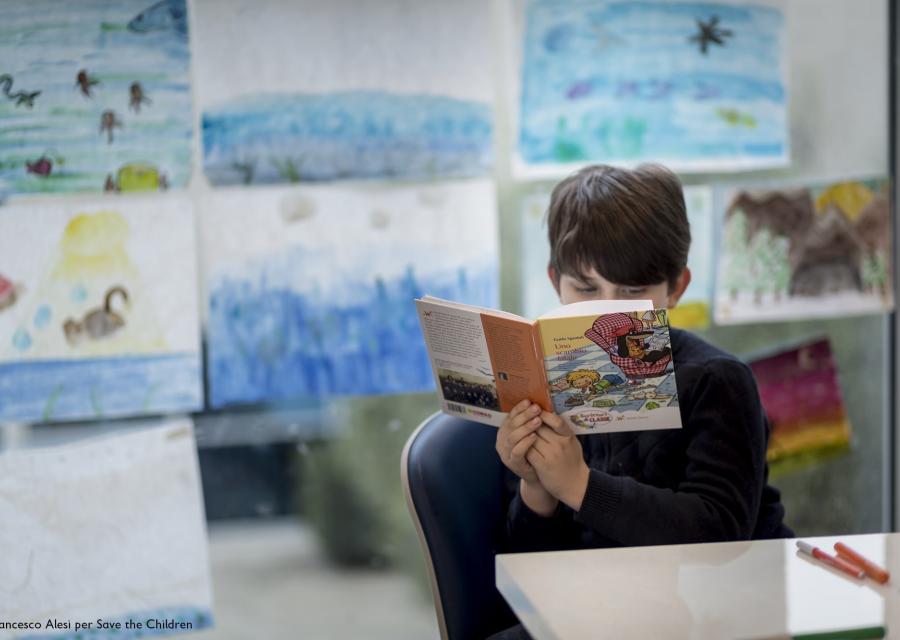 bambino seduto su una sedia davanti a un banco di scuola mentre legge un libro che tiene di fronte al suo viso