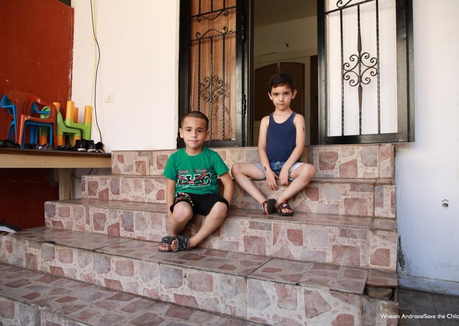 due bambini sono seduti sugli scalini di una casa, indossano abiti estivi. Uno ha la maglietta verde l altro blu