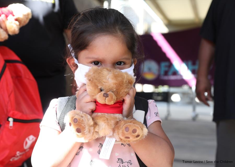 bimba con capelli castano scuro, bianca di carnagione, indossa una mascherina per proteggersi dal covid-19 e tiene tra le mani di fronte al viso un peluche a forma di orsacchiotto