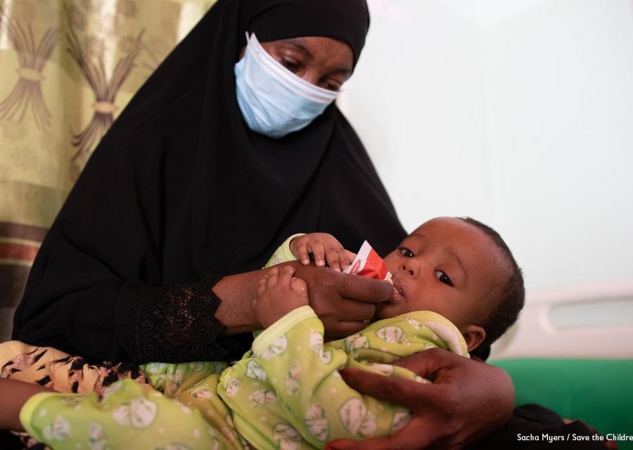 Una donna somala con tunica nera indossa mascherina ed è seduta su un letto. Sulle sue gambe tiene il figlio che ha una tutina gialla e lo imbocca facendogli mangiare cibo terapeutico.
