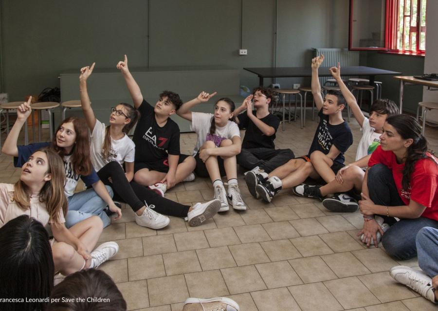 bambini e bambine della scuola primaria seduti a terra in cerchio che alzano la mano 