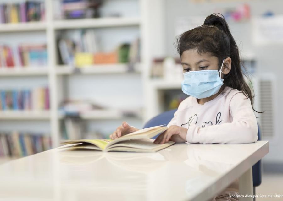 Bambina mora con coda ai lunghi capelli indossa mascherina chirurgica sul volto e una maglietta rosata legge un libro seduta a un tavolino