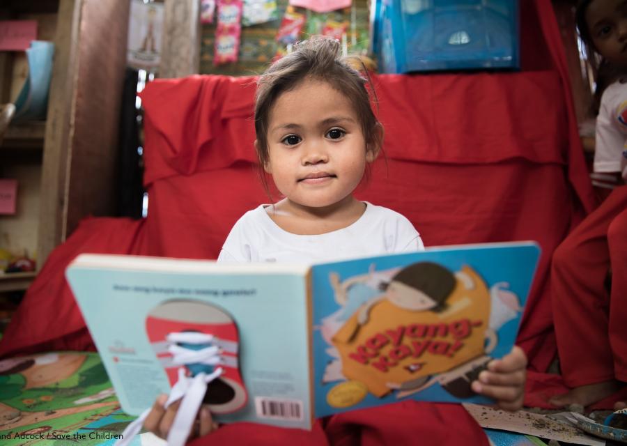 bimba del sud est asiatico con occhi scuri e capelli castani seduta con un libro grande quanto lei aperto innanzi a sè. 