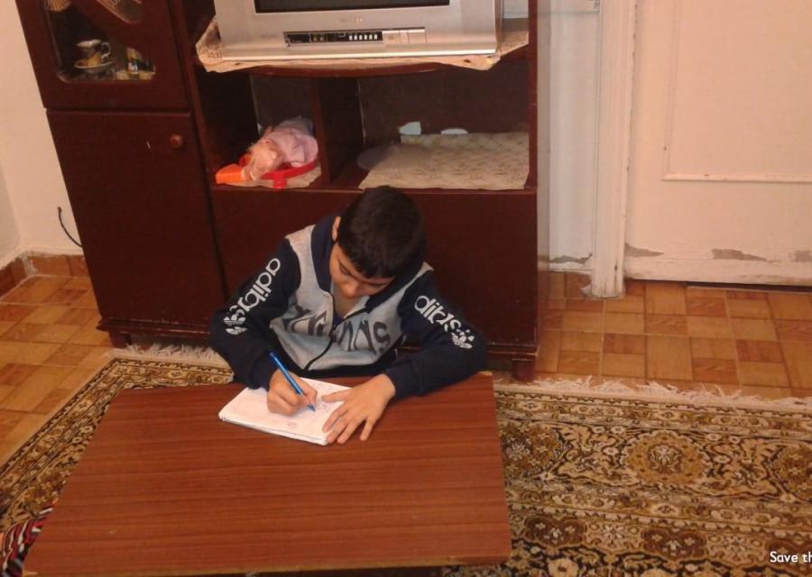 Bambino libanese di circa 12 anni è seduto a terra su un tappeto e scrive su un figlio bianco appoggiato a una tavolo di legno.