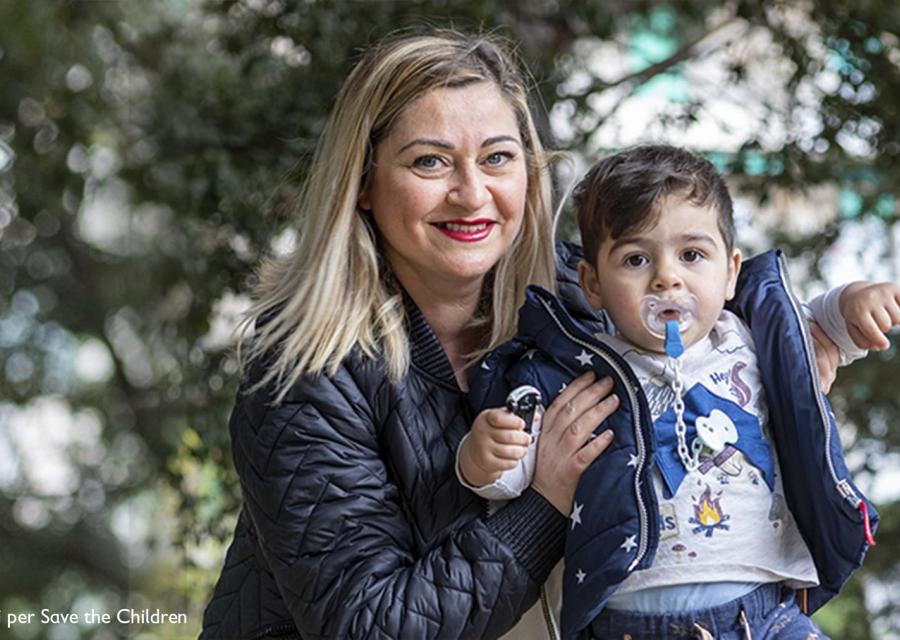 Una mamma bionda con capelli fino alle spalle è molto sorridente e ha occhi chiari, mantiene in piedi suo figlio di circa un anno con il ciuccio in bocca.