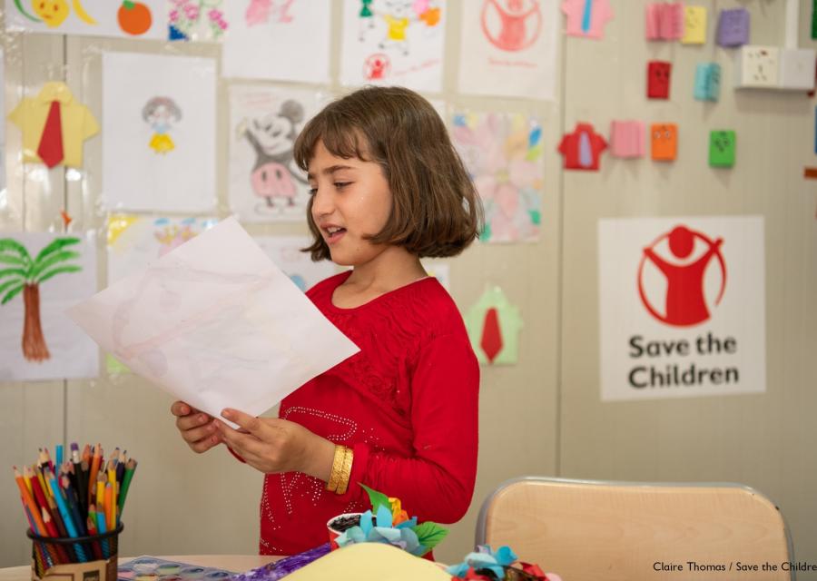 bambina in classe, in piedi, mentre legge ad alta voce qualcosa da un foglio. Sullo sfondo la parete dell aula è ghermita di disegni dei bambini e delle bambine.