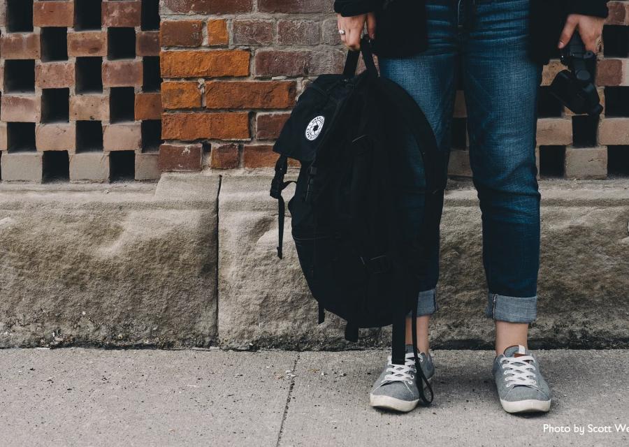 gambe di una studentessa che tiene in mano uno zaino nero. Sullo sfondo muro di mattoncini