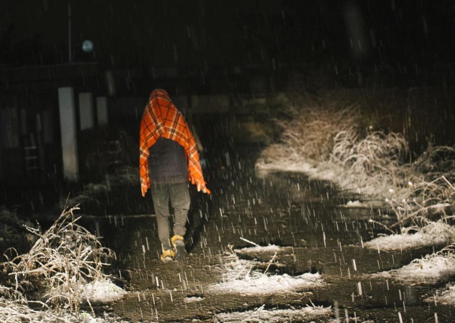 Foto notturna in un viale oscuro mentre sta nevicando. Si vede di spalle un ragazzo che cammina lungo il viale e sotto la neve con pantaloni scuri e una coperta arancione posta sopra le spalle e la testa.