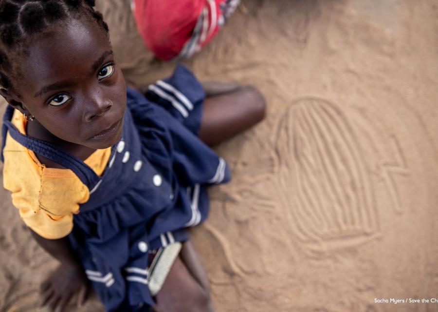 Bambina mozambicana ripresa dall alto è seduta a terra e guarda verso l alto. Indossa un vestito blu e una maglietta arancione.