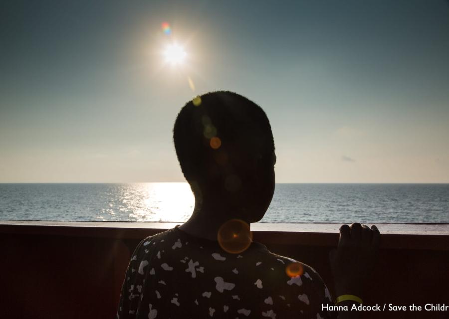 Un bambino contro sole di spalle è affacciato alla balaustra di una nave e guarda il mare