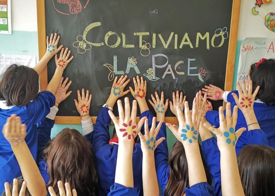 bambine e bambini a scuola, con divisa blu, di spalle alzano le braccia verso la lavagna. Sulle mani tanti fiori disegnati e sulla lavagna la scritta coltiviamo la pace.