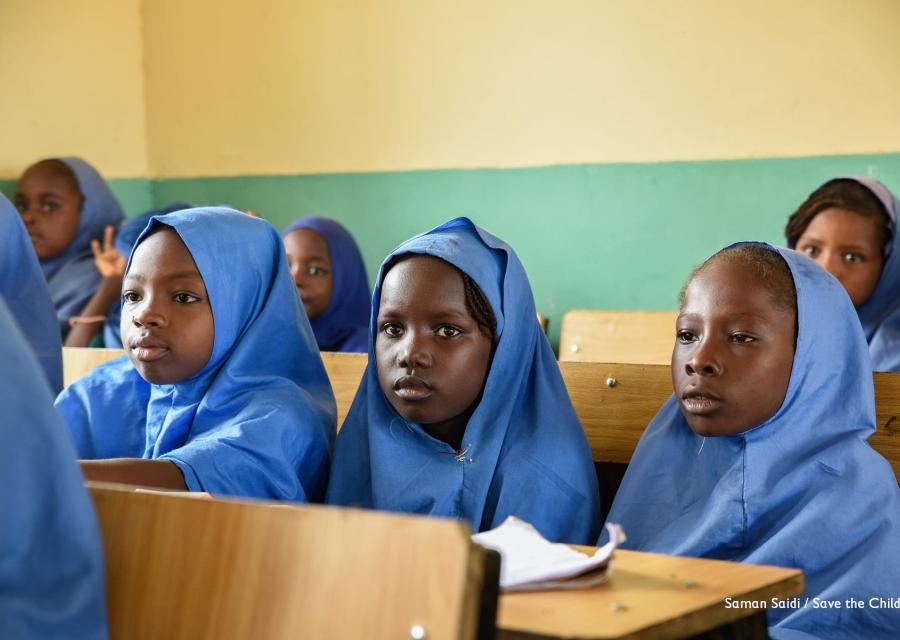 Tre bambine nigeriane della scuola primaria sono sedute a banchi scolastici in legno e guardano davanti a loro. Sono vestite con una divisa celeste e il loro capo è coperto.