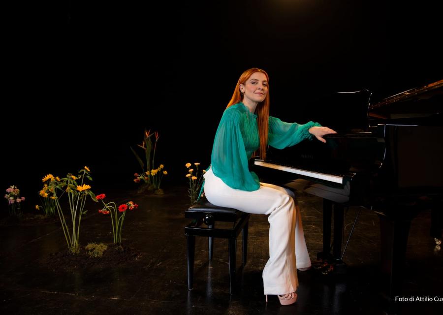 La cantante Noemi è seduta a un pianoforte su un set con alcuni fiori finti alle spalle. Indossa una camicia verde acqua e pantaloni bianchi.