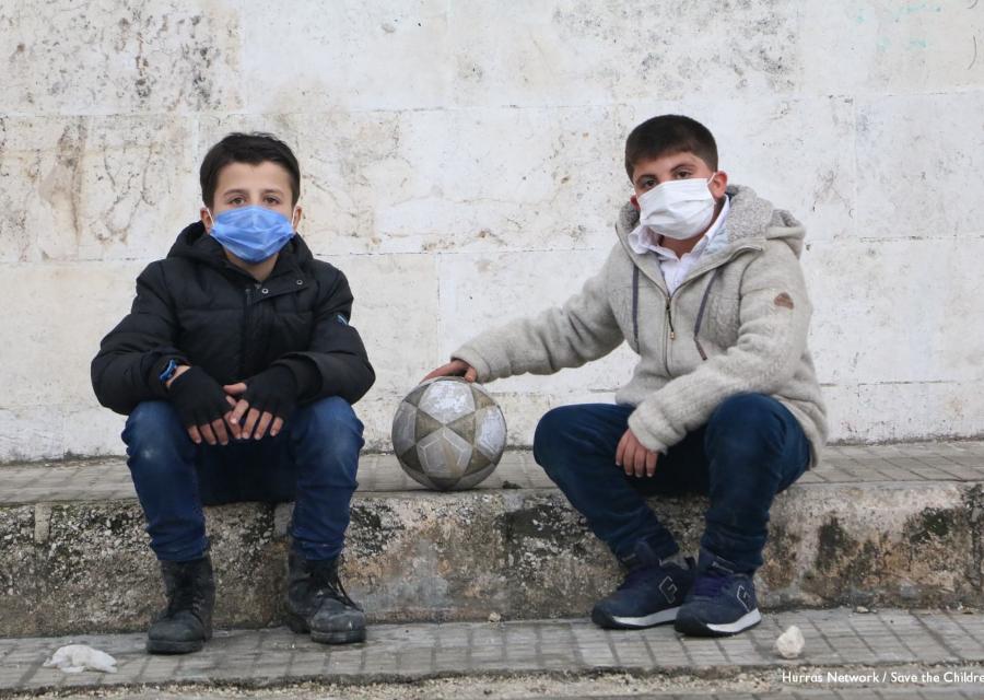 Due bambini siriani di 10 anni indossano la mascherina, un cappotto e dei jeans e sono seduti uno a fianco all altro su un gradino. Uno dei due tiene il braccio allungato lateralmente con la mano posata su un pallone, poggiato anch esso sul gradino.