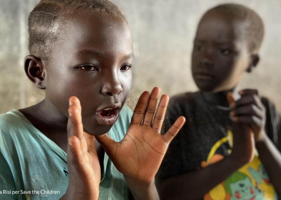 Un bambino nero sullo sfondo batte le mani mentre un bambino nero in primo piano fa un gesto con la mano poggiando i pollici sotto il mento con la mano aperta