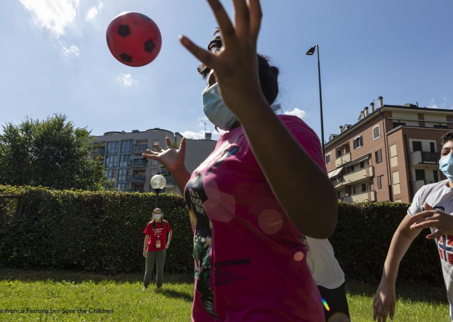 in primo piano una ragazza che gioca con a pallone con altri 2 ragazzi sullo sfondo e una operatrice save the children. Giocano all aperto su un campo verde e indossano la mascherina al volto.