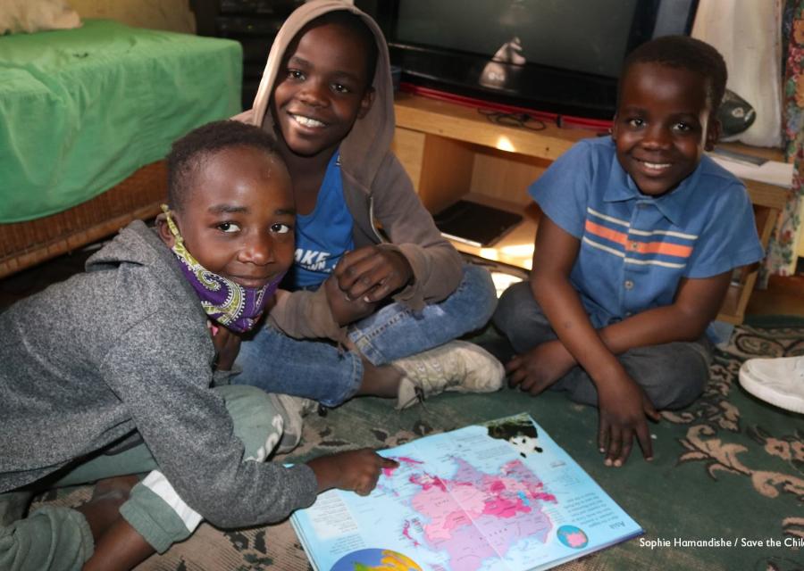 Tre bambini dello Zimbabwe sono seduti a terra intorno a un libro insieme. Hanno gli sguardi sorridenti e guardano in camera