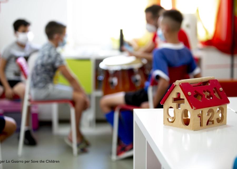 3 bambini sfocati sullo sfondo seduti a un tavolino con le mascherine e un adulto, operatore save the children, gli spiega qualcosa. In primo piano una casa giocattolo appoggiata su un tavolo.