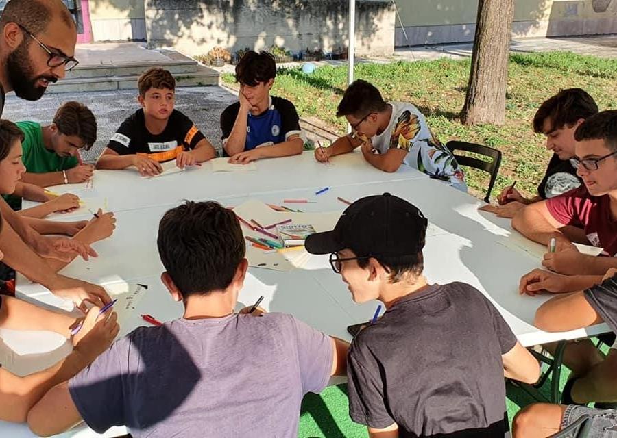 Studenti intorno a una tavola rotonda scrivono e studiano, un adulto li supervisiona