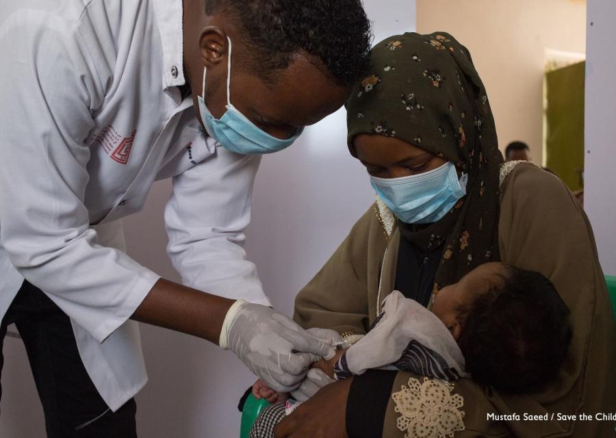 Un operatore save the children con indosso camice e mascherina vaccina un neonato in braccio a sua madre anche lei con maschrina