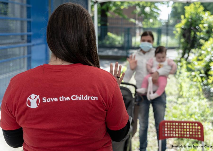 operatrice di save the children di spalle che saluta una mamma con un bimbo in braccio