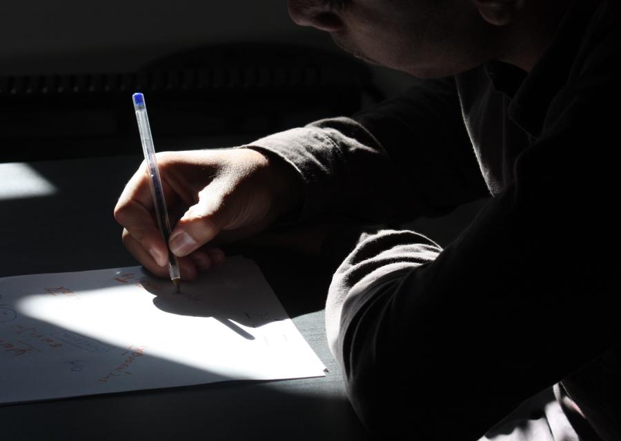 Braccia di un bambino poggiate su un tavolo, una mano scrive con una penna su un foglio bianco. Tutta la foto è in penombra 